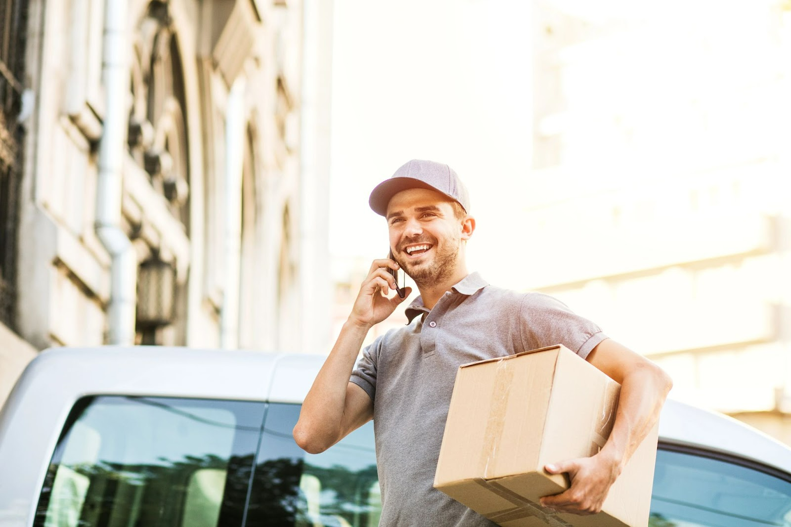 Smiling delivery man holding a cardboard box and talking on the phone in front of a van.