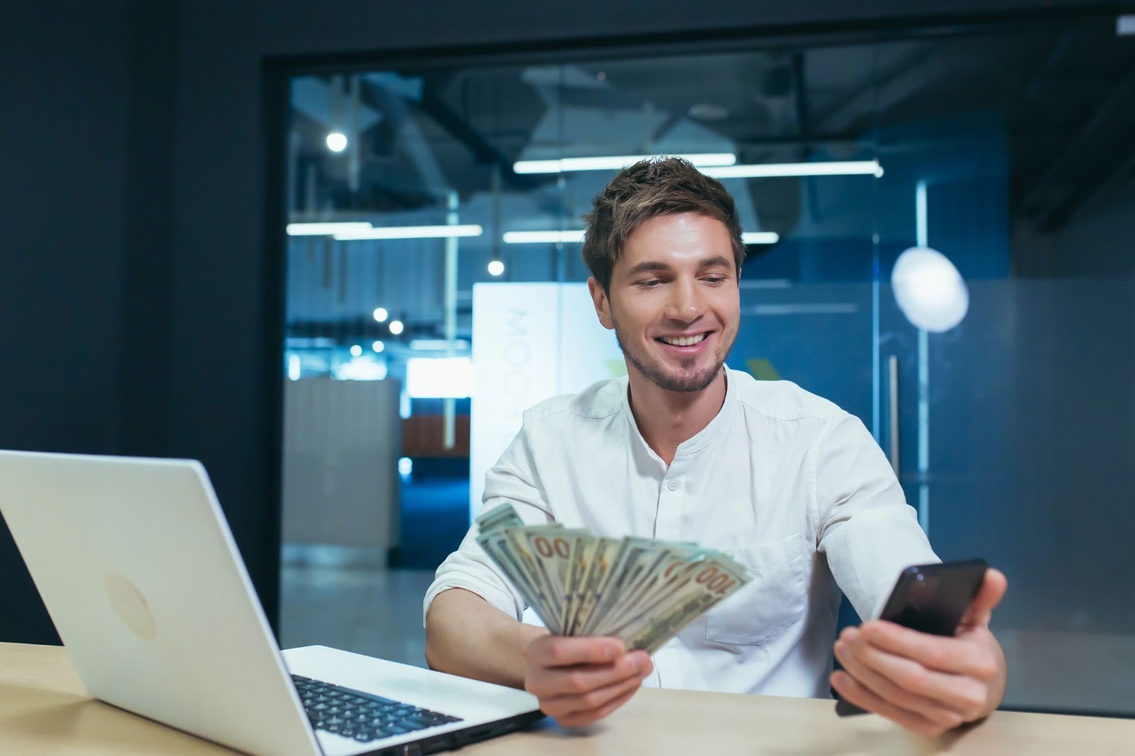 Freelancer smiling while holding cash and checking a smartphone, sitting at a desk with a laptop.