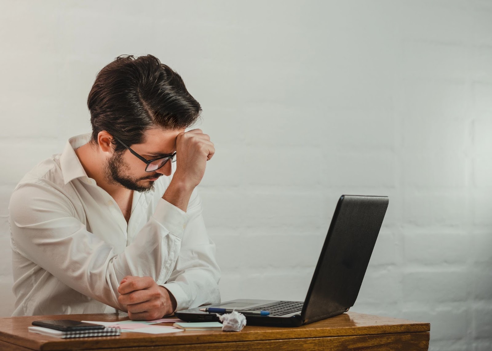 Stressed man sitting at a desk with a laptop, holding his head while looking down at paperwork