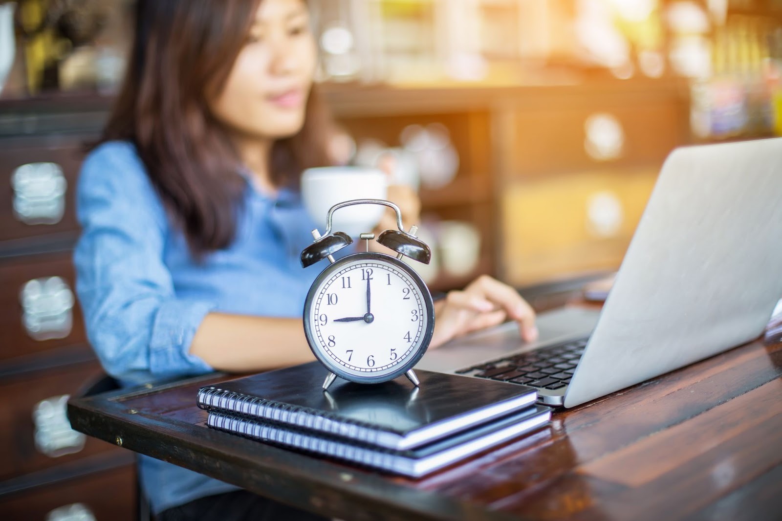 Alarm clock on a desk in front of a woman working on a laptop.