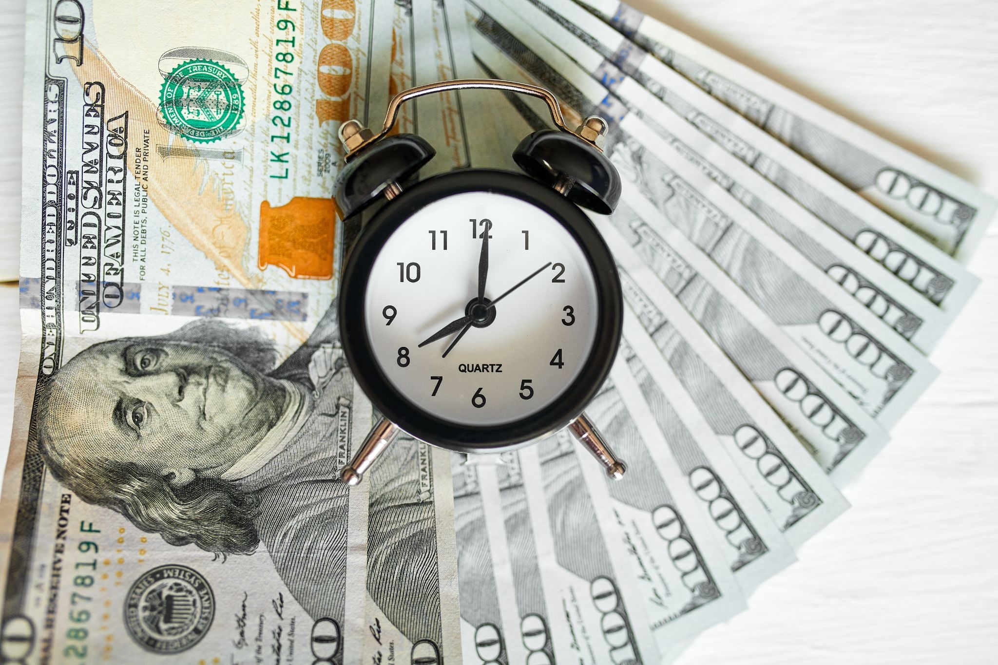 A black vintage alarm clock placed on top of fanned-out US $100 bills on a white surface, symbolizing the concept of time and money.