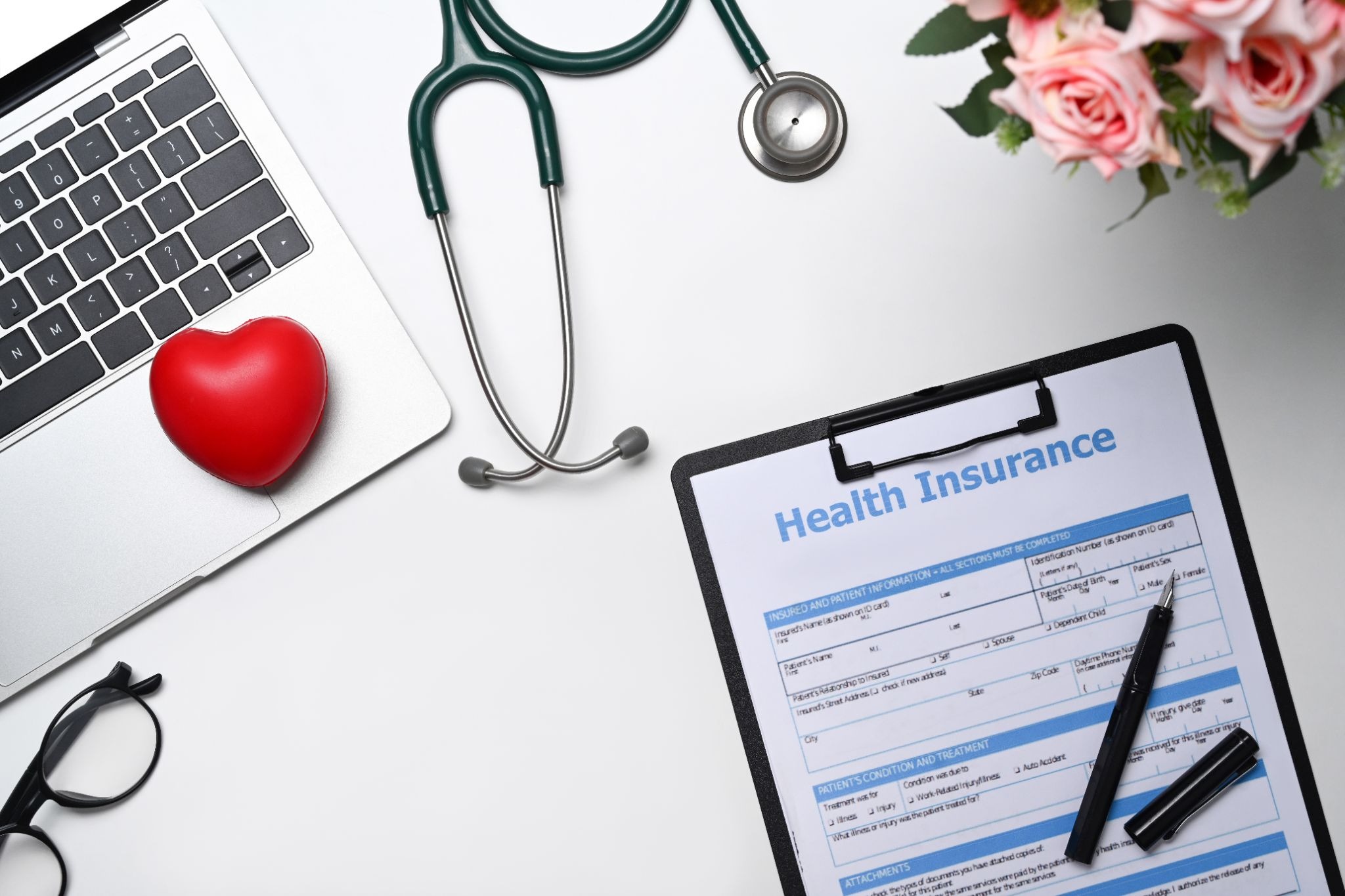 Flat lay of a health insurance form on a clipboard with a pen, a stethoscope, laptop, red heart, eyeglasses, and pink flowers on a white surface.