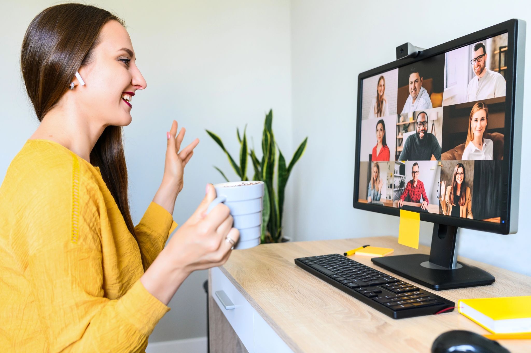 Woman holding a coffee mug and smiling while participating in a video call with multiple people displayed on her computer screen.