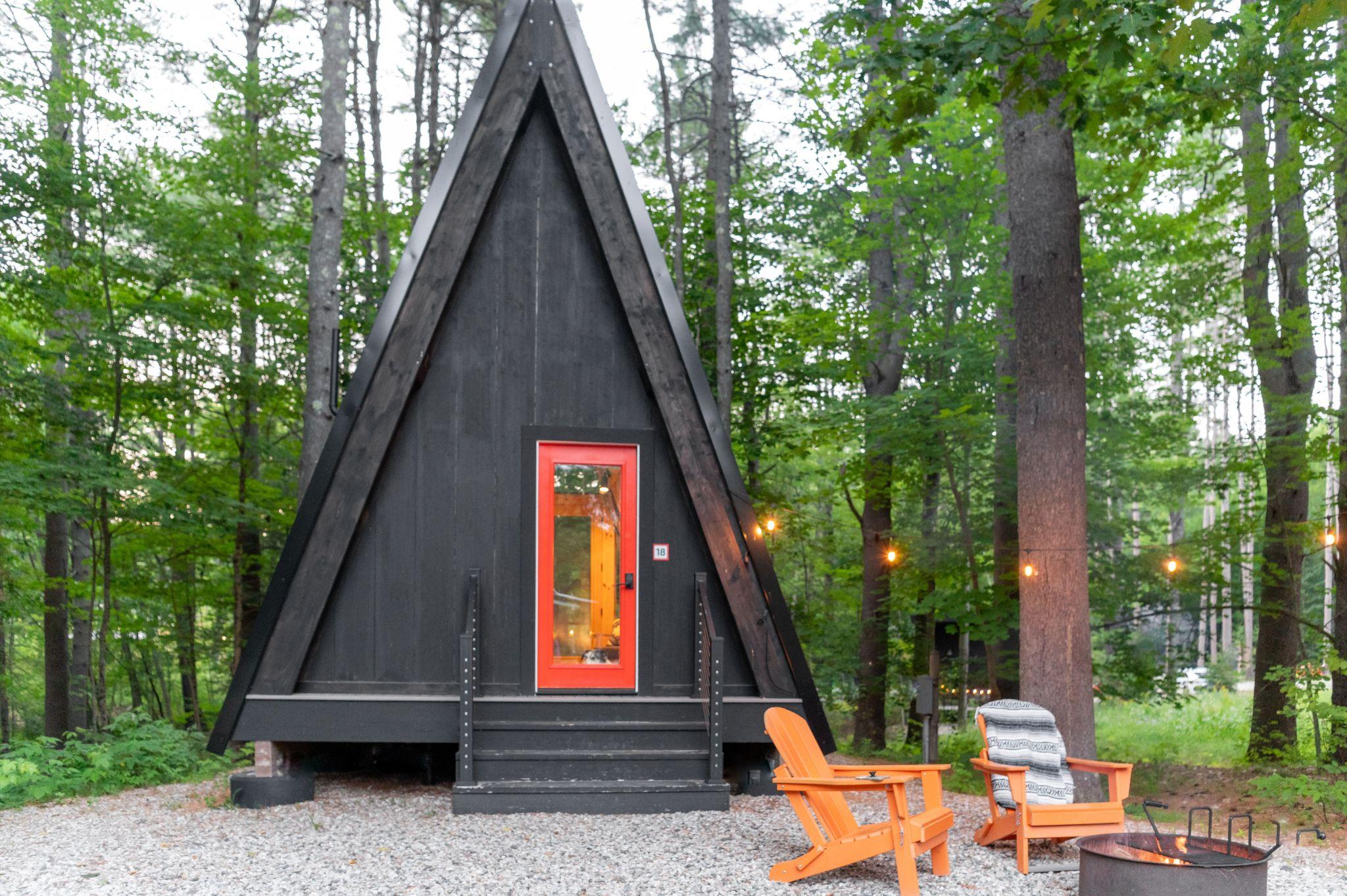A small cabin surrounded by trees, featuring dark black walls, a bright red door, and two chairs
