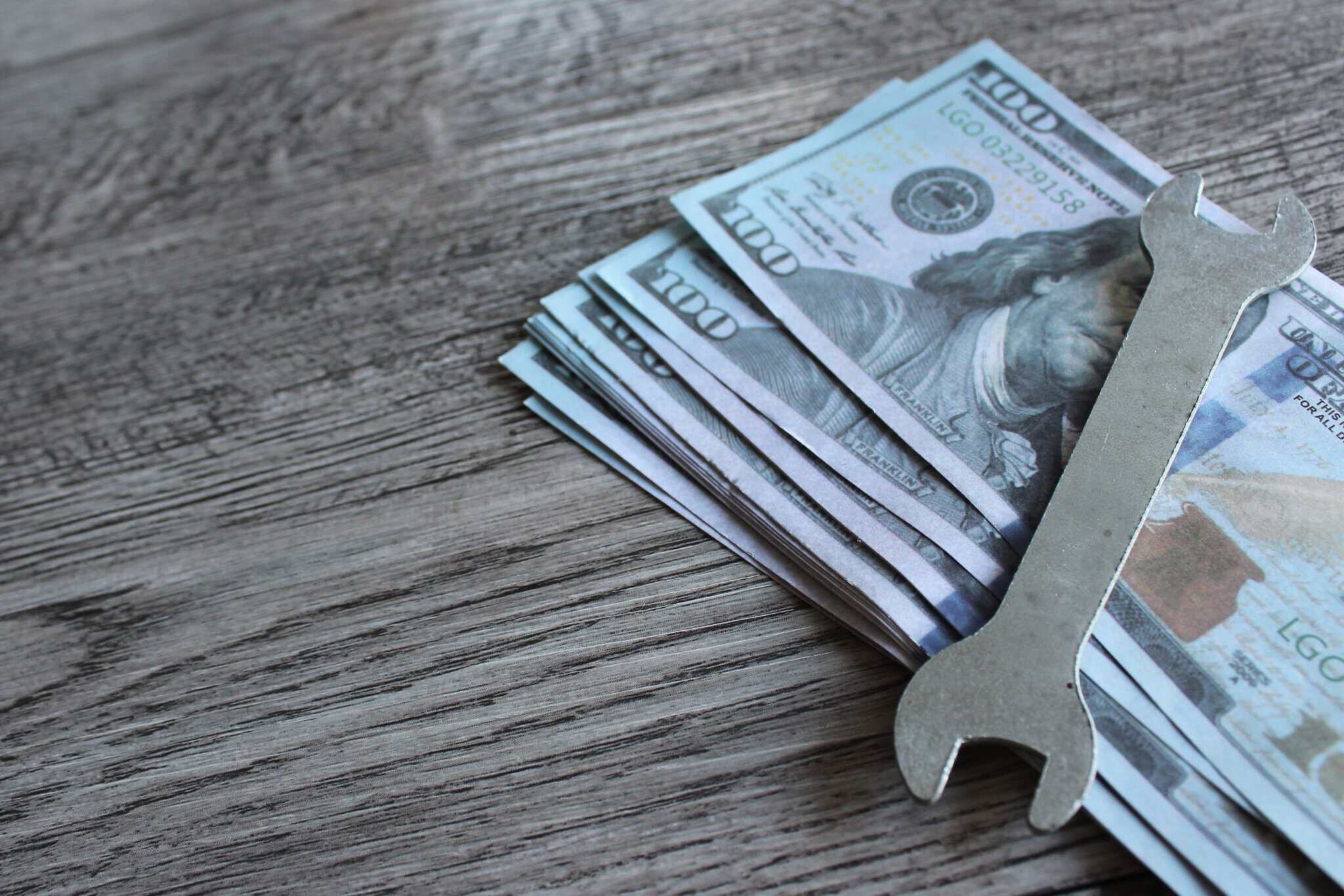 Stack of $100 bills with a wrench on a wooden table, representing planning for maintenance and repair costs.